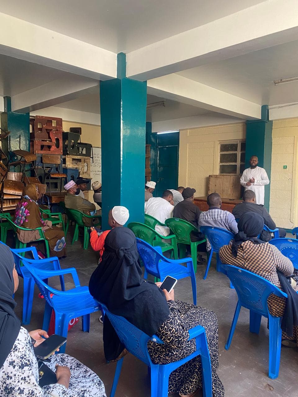 A man in a white kanzu speaking to members seated on green and blue plastic chairs in a community hall.