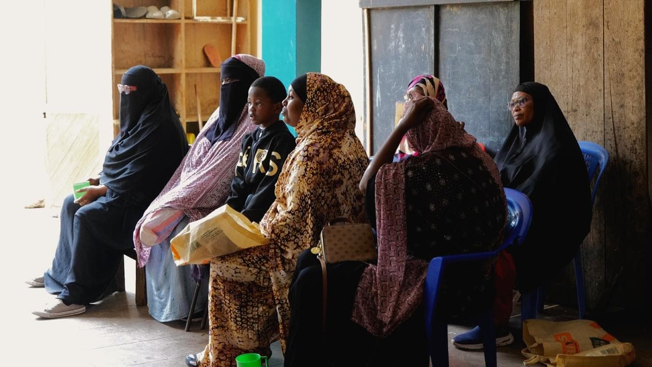 Women in hijab and a boy seated indoors on a bench and blue chairs, with bags on their laps.