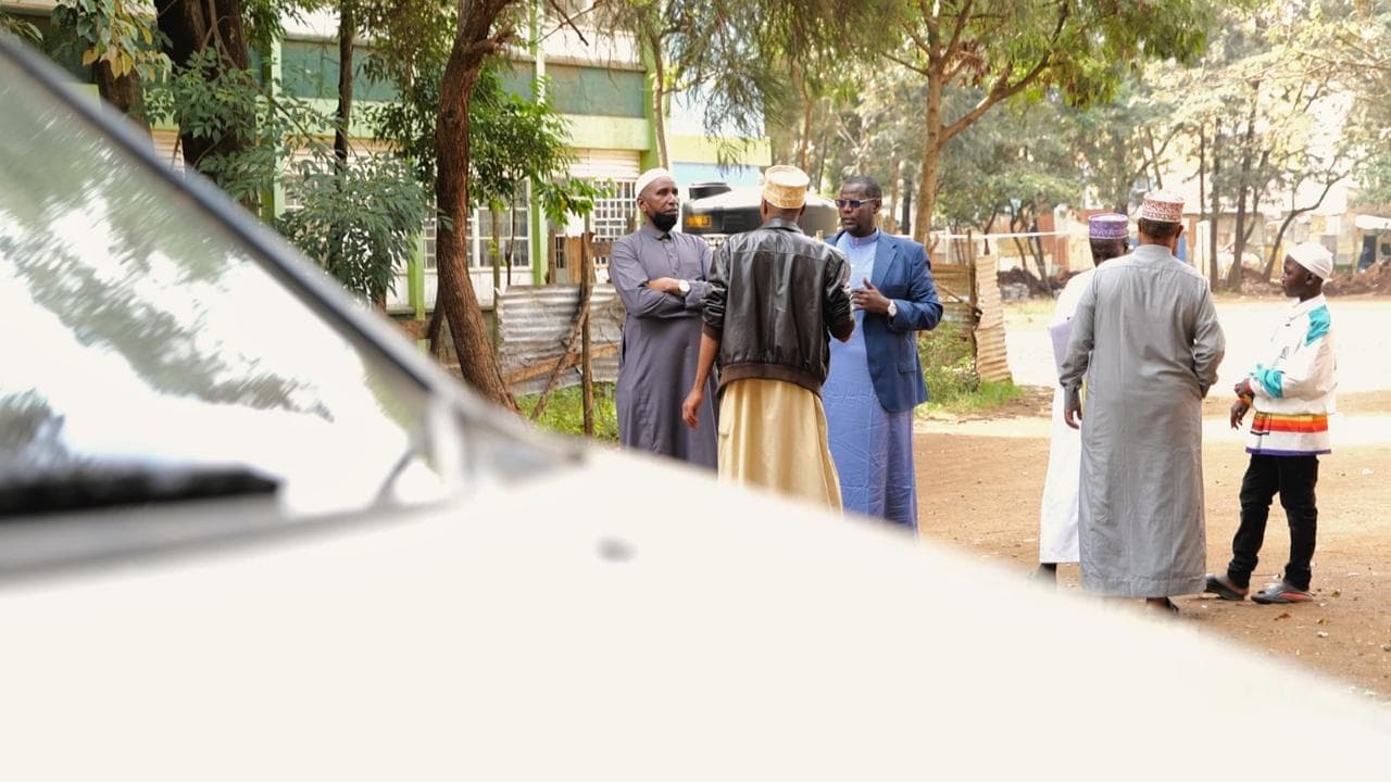 Men in kanzus and kufis talking on a dirt path under trees, with the bonnet of a white car in the foreground.