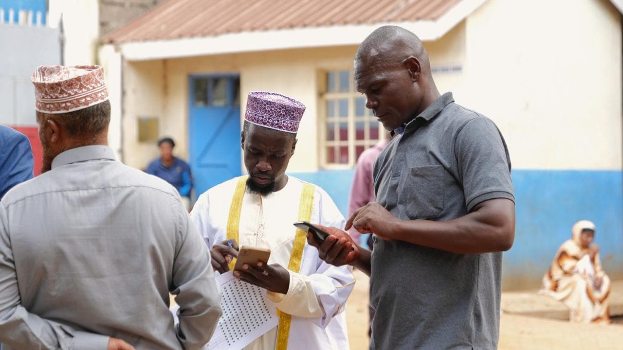 Three men outside a building, looking at phones and a printed list of dotted rows.